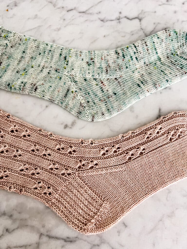 A close-up on the heels of two handknit socks laid out flat on a white marble countertop. The pink sock, on the bottom, has a heel flap and gusset. The green sock, on the top, has a Fleegle heel.