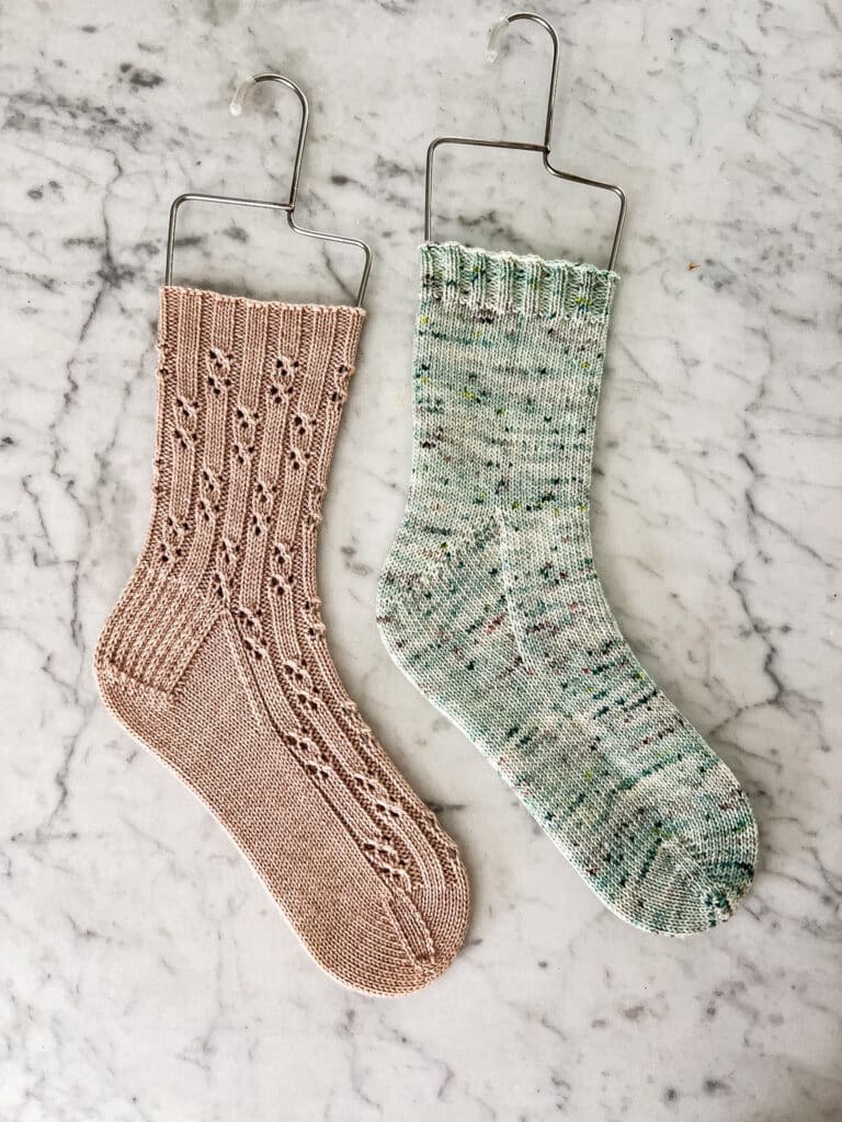 A top-down photo of two handknit socks displayed on wire sock blockers on a white marble countertop. The pink sock, on the left, has a heel flap and gusset. The green sock, on the right, has a Fleegle heel.