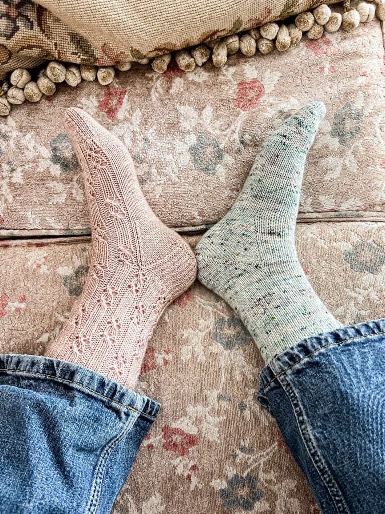 A woman's feet wearing two mismatched socks rest on the faded floral cushions of a vintage sofa. The left foot wears a pink sock with a heel flap and gusset. The right foot wears a green sock with a Fleegle heel.