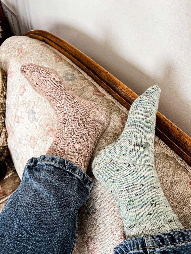A woman's feet wearing mismatched handknit socks rest on the back of a vintage sofa with faded floral upholstery. The left foot wears a pink sock with a heel flap and gusset. The right foot wears a green sock with a Fleegle heel.