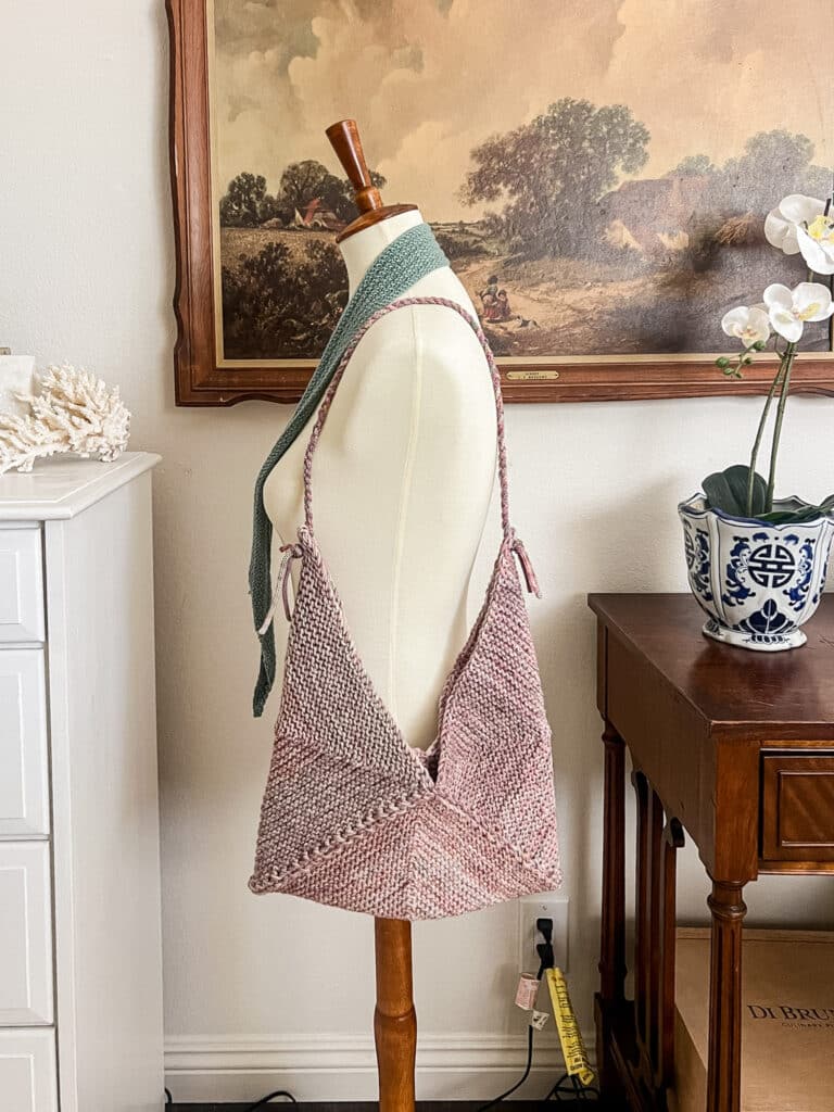 A straight-on photo of a pink knit bag made from mitered squares that's hanging from the shoulder of a white dressmaker's form.