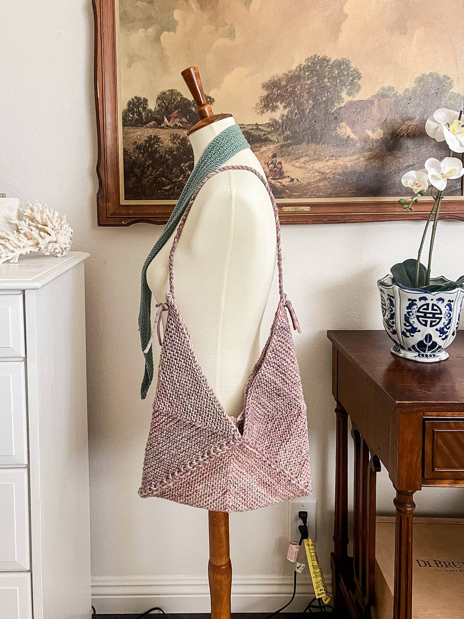 A straight-on photo of a pink knit bag made from mitered squares that's hanging from the shoulder of a white dressmaker's form.