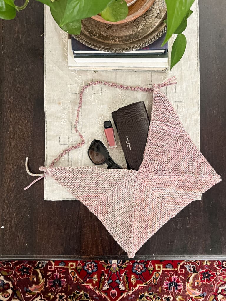 A pink knit bag made from mitered squares is laid out on a tabletop with a wallet, sunglasses, and lip gloss poking out from the top.