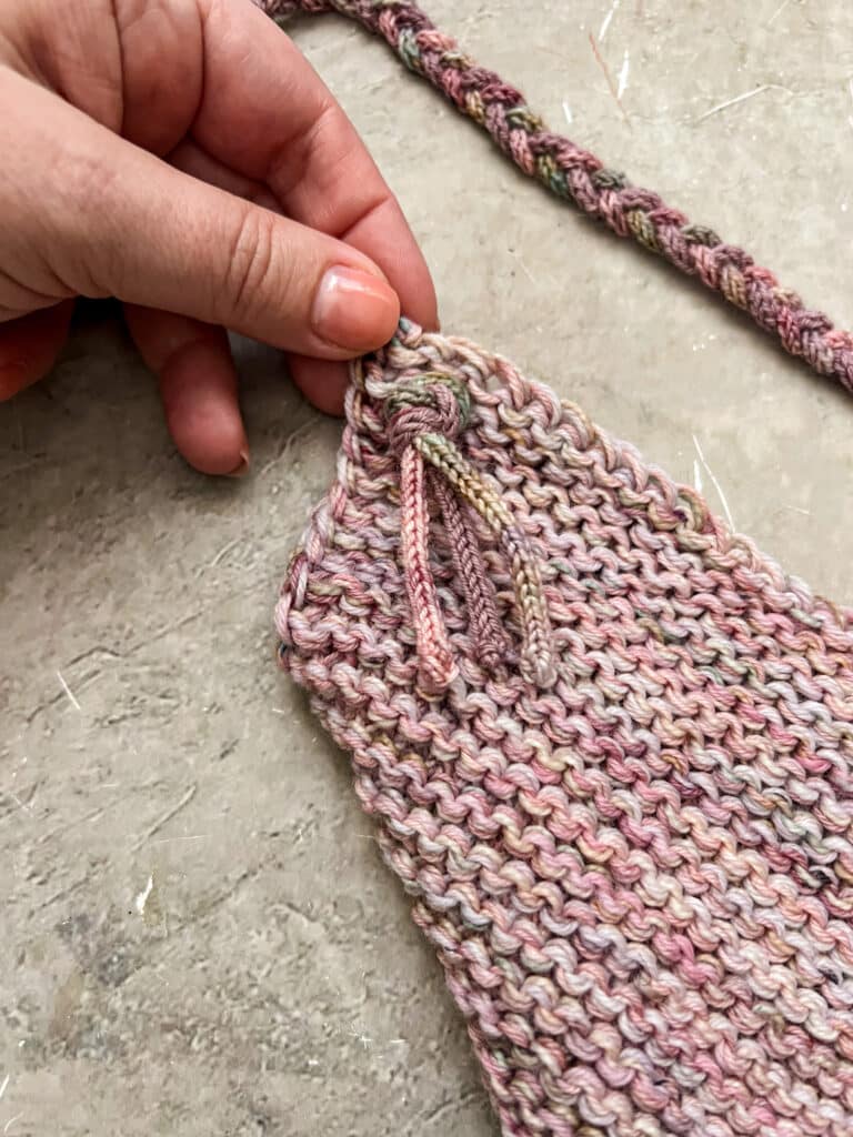 A white woman's left hand holds the corner of a knit mitered square. Three I-cords braided together have been pushed through the fabric and tied off with a large knot.