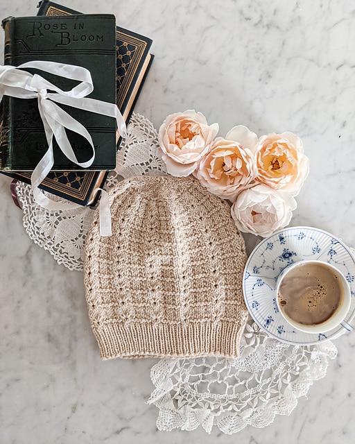 A peach-colored handknit hat lays flat on a white marble countertop. The stitch pattern is created with eyelets, purl stitches, and decreases.