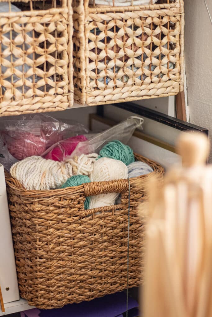 A square woven basket filled with lots of different colors of yarn sits on a shelf surrounded by books and papers.
