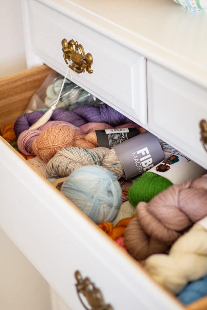 A drawer on a white dresser is pulled out to show lots of different colors of fingering-weight yarn in hanks and skeins.
