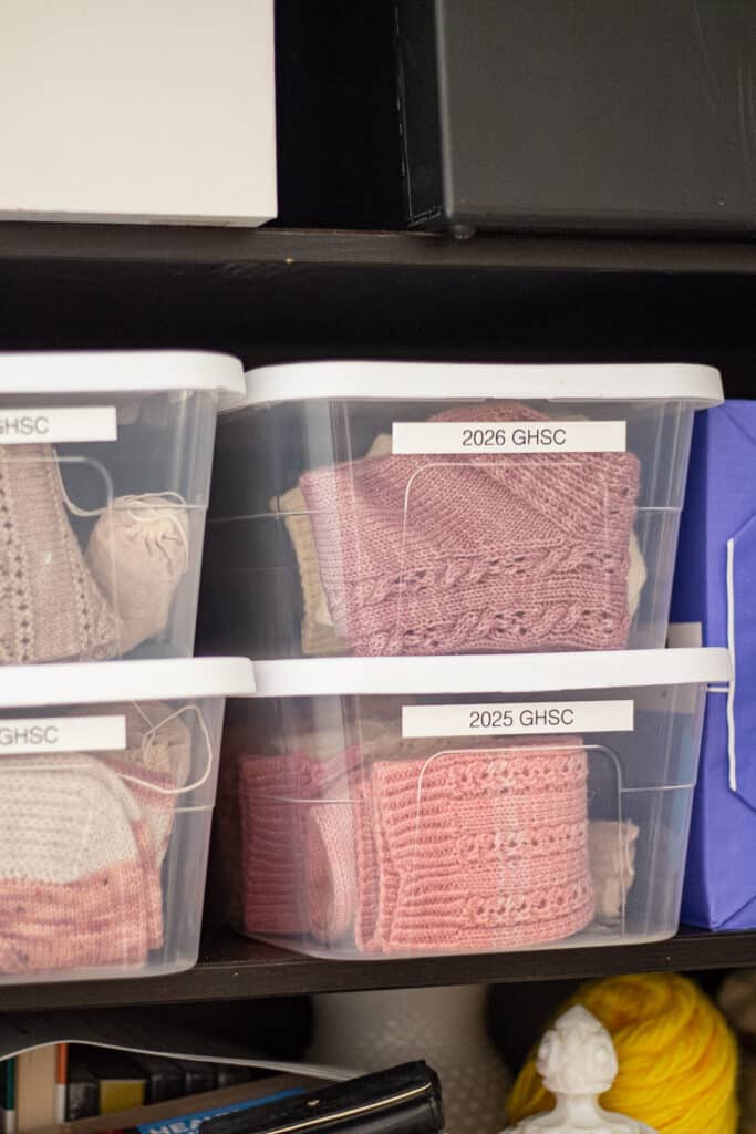 Four clear, plastic tubs with white lids sit on a shelf in a dark bookcase. Inside the bins are folded, handknit socks.