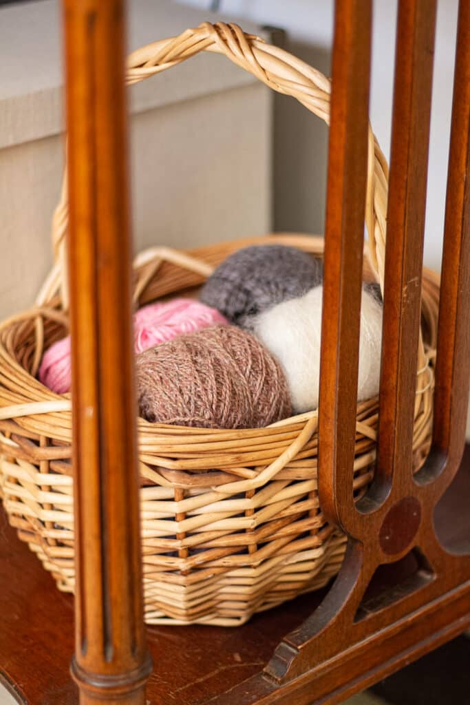 A light brown wicker basket filled with lots of remnants of yarn is photographed through the side legs of a table.