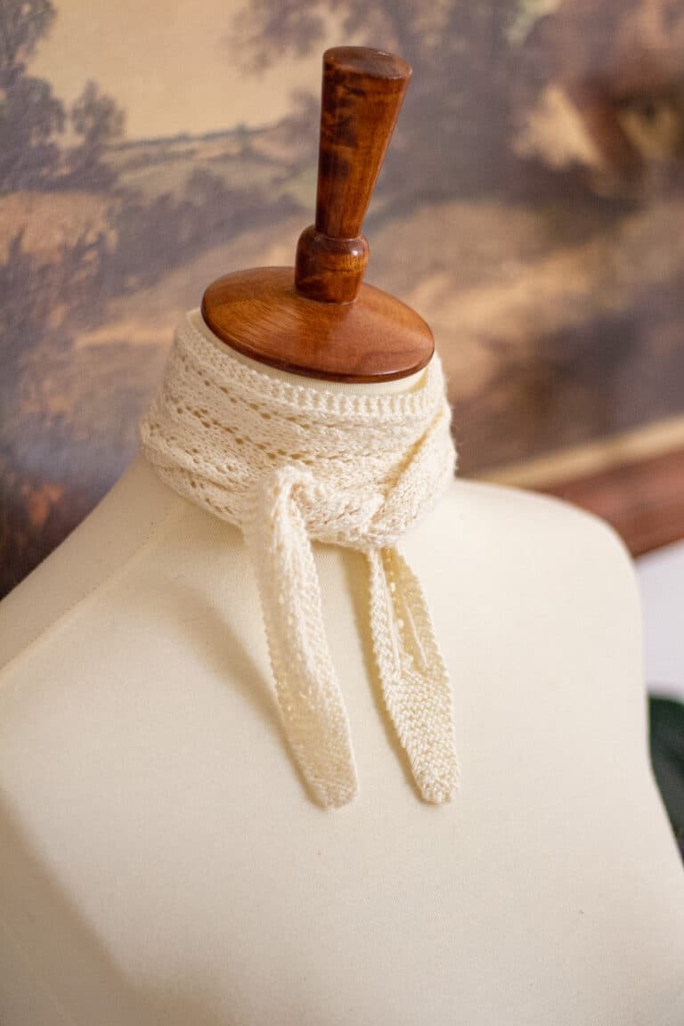 A white mini scarf is wrapped twice and tied around the neck of a white dressmaker's form.