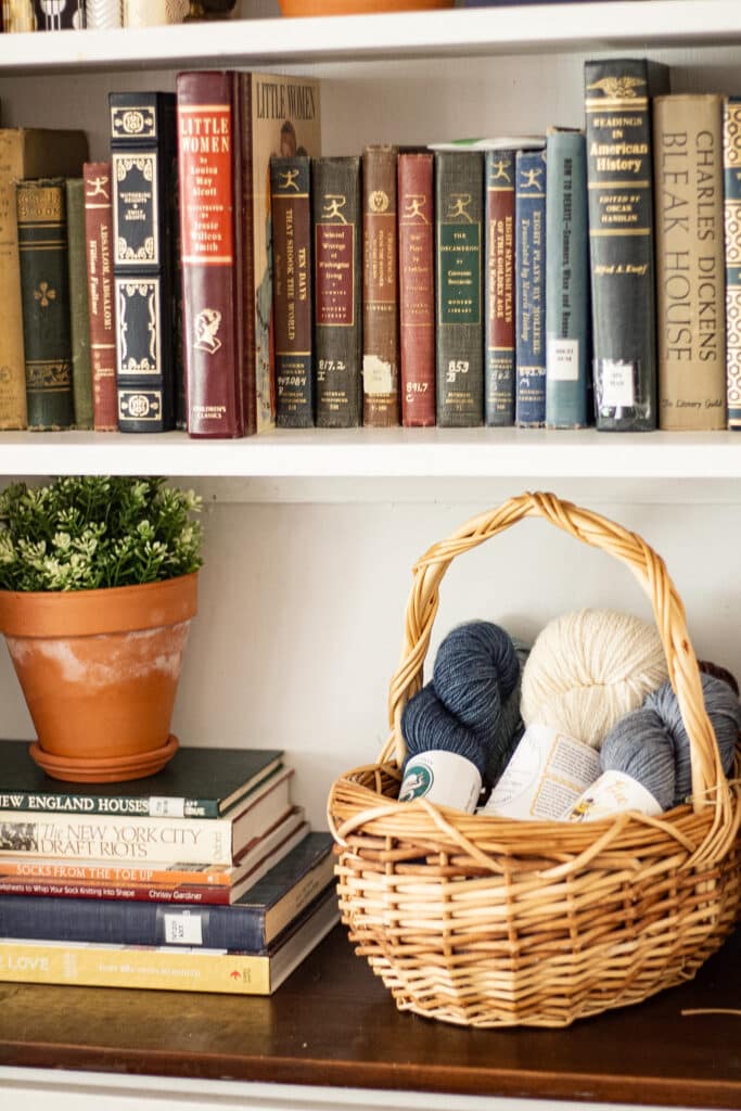 A wicker basket of blue and white hanks of yarn sits on a bookshelf surrounded by vintage and antique books and a potted plant.