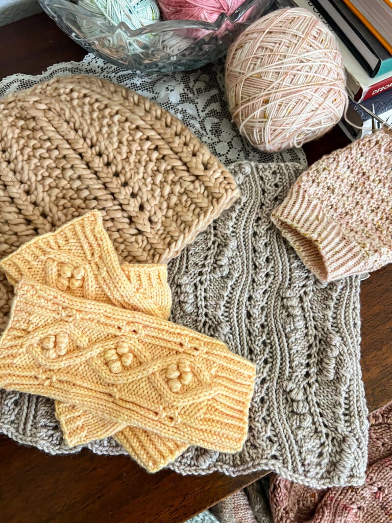A top down photo of several knit items in pink, gray, and yellow, all of them featuring twisted stitches as an intentional design element.