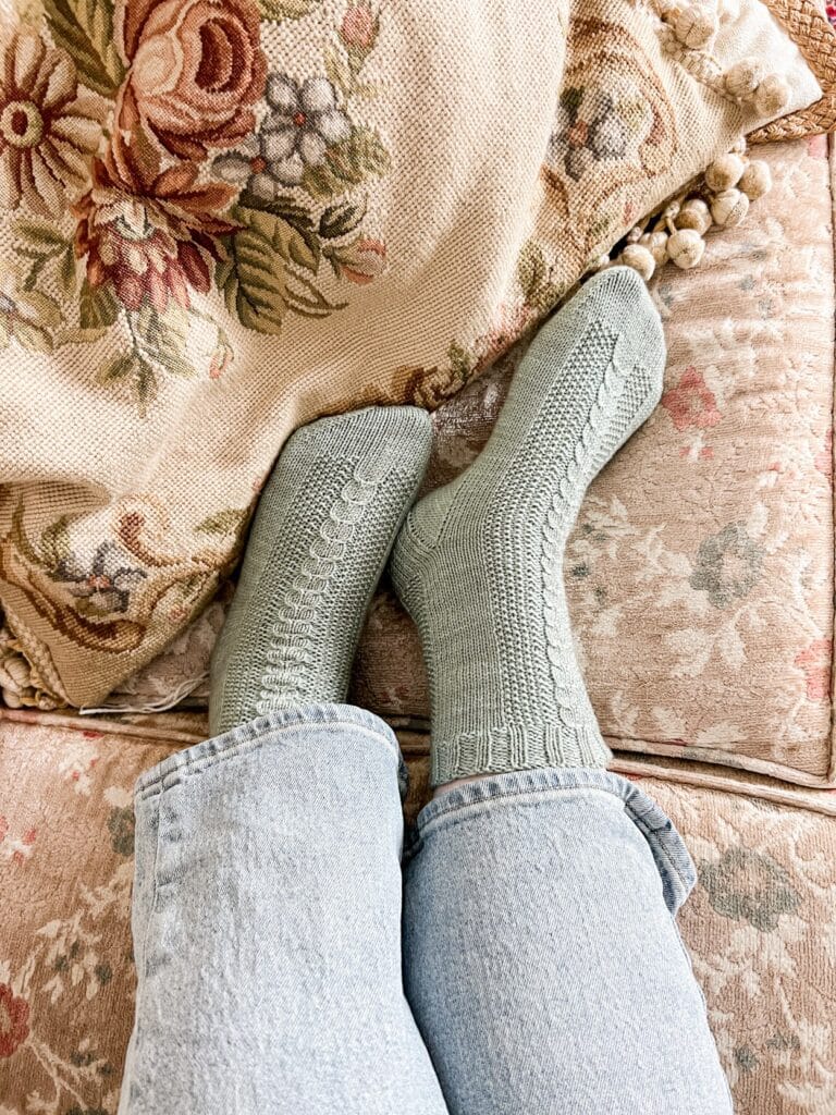 A woman's feet wearing a pair of light green handknit socks rest against a needlepoint cushion. The feet are pointing to the right.