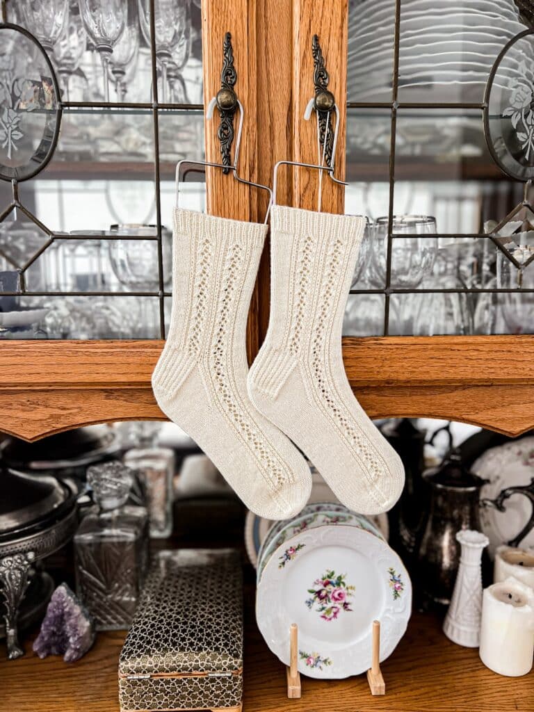A pair of white handknit socks with lacy side panels hangs on sock blockers from the doorknobs of an oak china hutch.