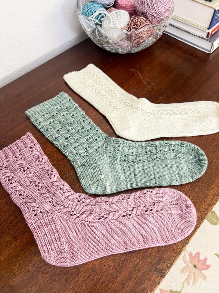 Three solo socks in shades of purple, green, and white sit on top of a dark wood table. The toes are all pointing to the right.