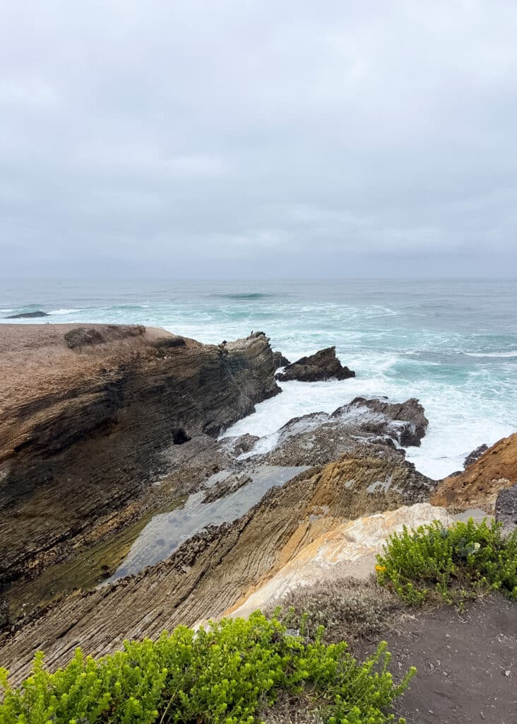 A view of a rocky part of the coastline with high surf and bright, aqua-colored water. The sky is very cloudy.