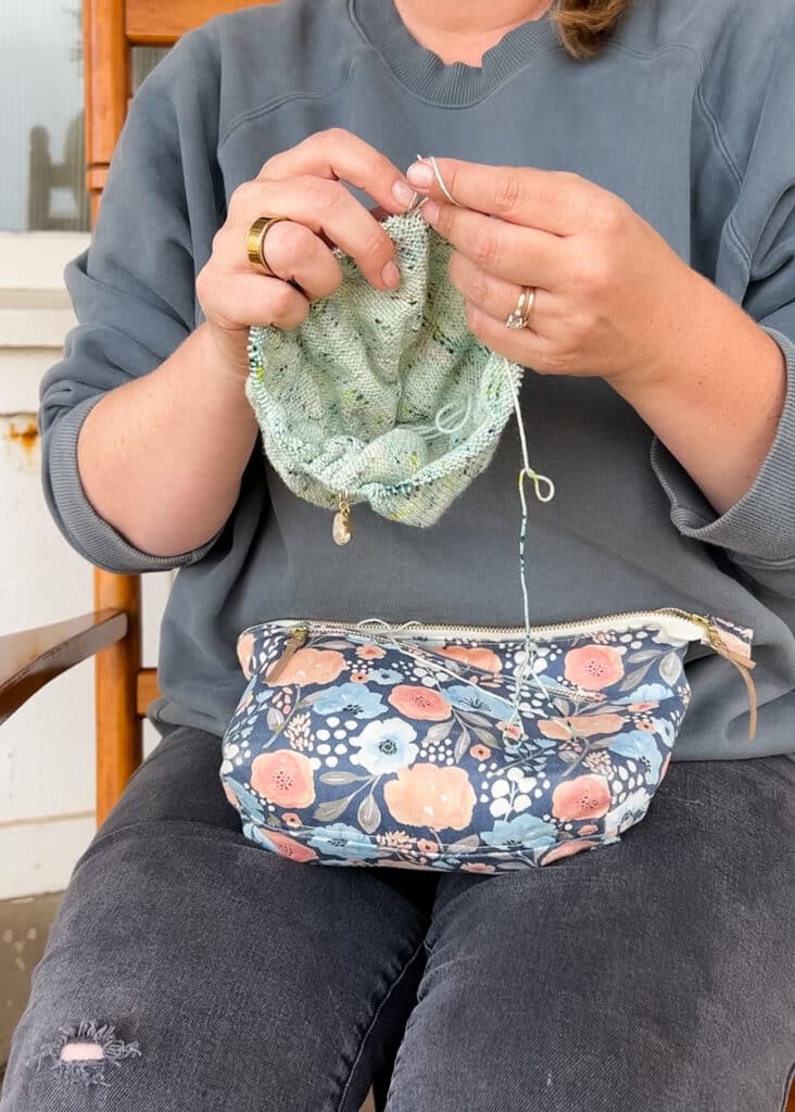 A white woman in a dusty blue sweatshirt and dusty gray jeans with a hole in one leg knits a light green stockinette project.
