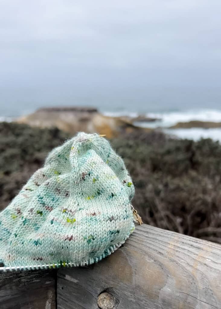 A green stockinette knitting project sits on a wooden fence rail on a bluff overlooking a rocky beach with high surf.