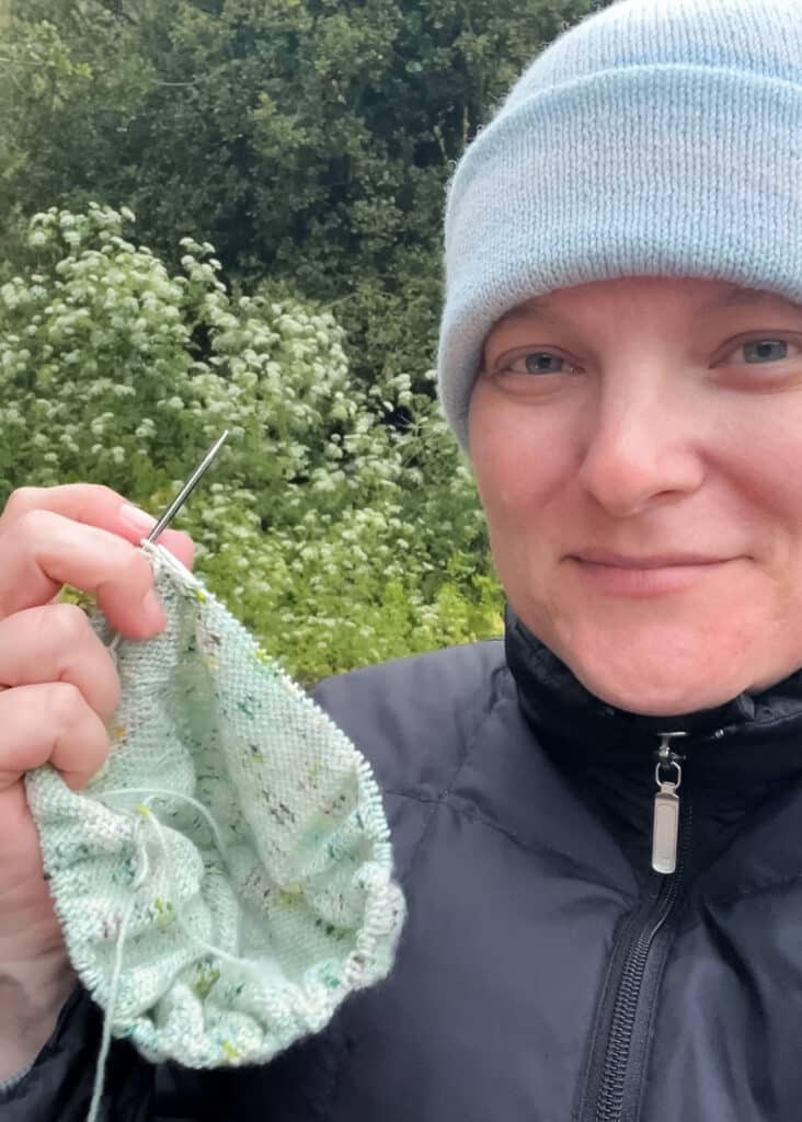 A white woman in a light blue knit cap smiles softly while holding up her green stockinette knitting project.