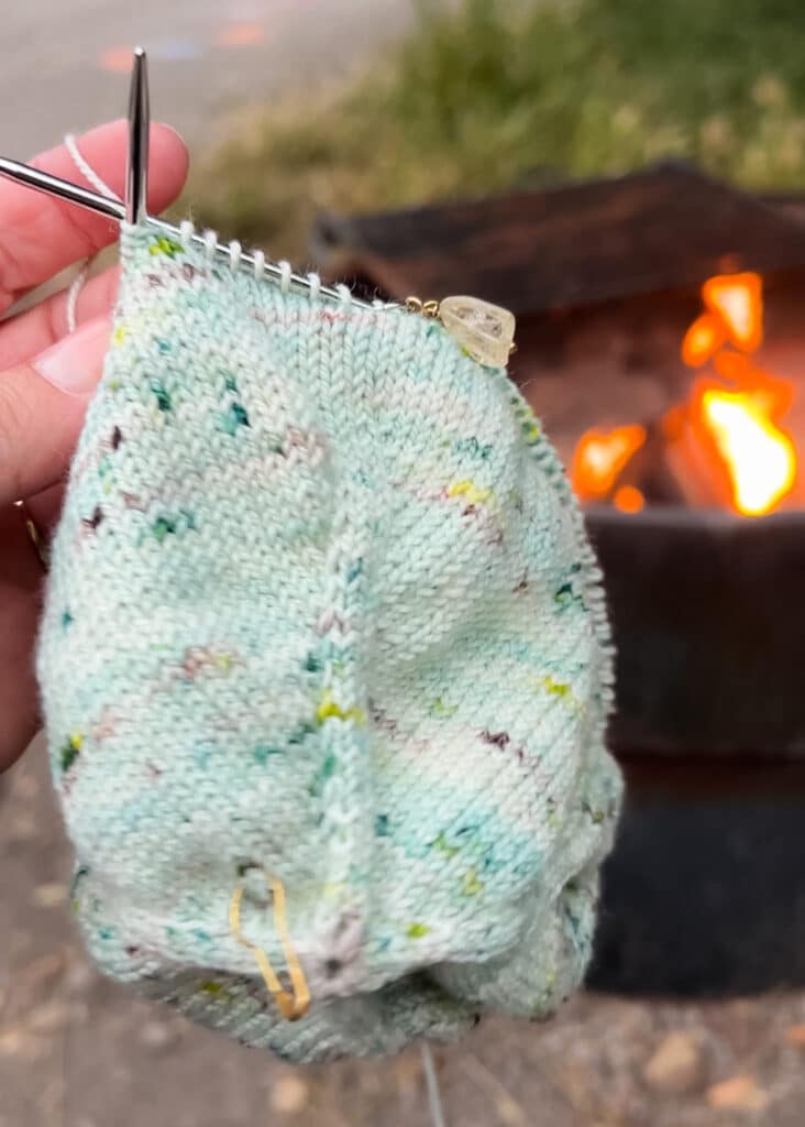 A white woman's left hand holds up a bit of green stockinette knitting in front of a campfire.