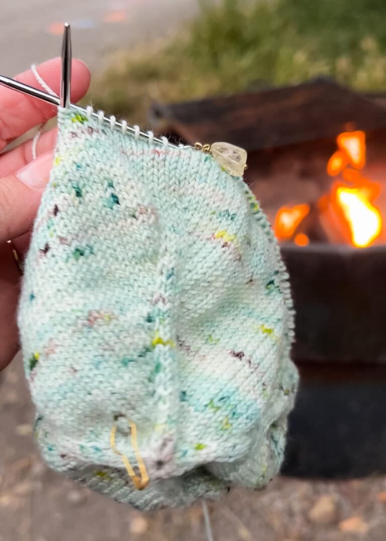 A white woman's left hand holds up a bit of green stockinette knitting in front of a campfire.