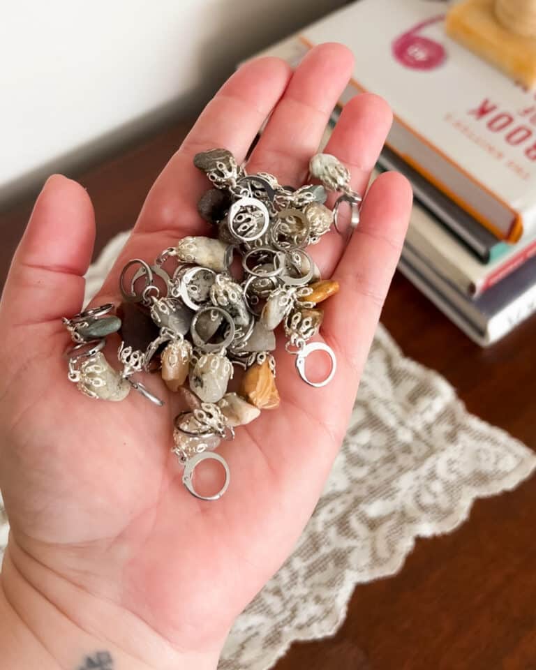 A white woman's left hand holds a jumble of many stitch markers made from beach pebbles.