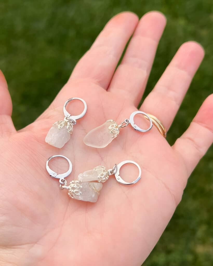 A white woman's left hand holds four stitch markers made from white sea glass and beach pebbles.