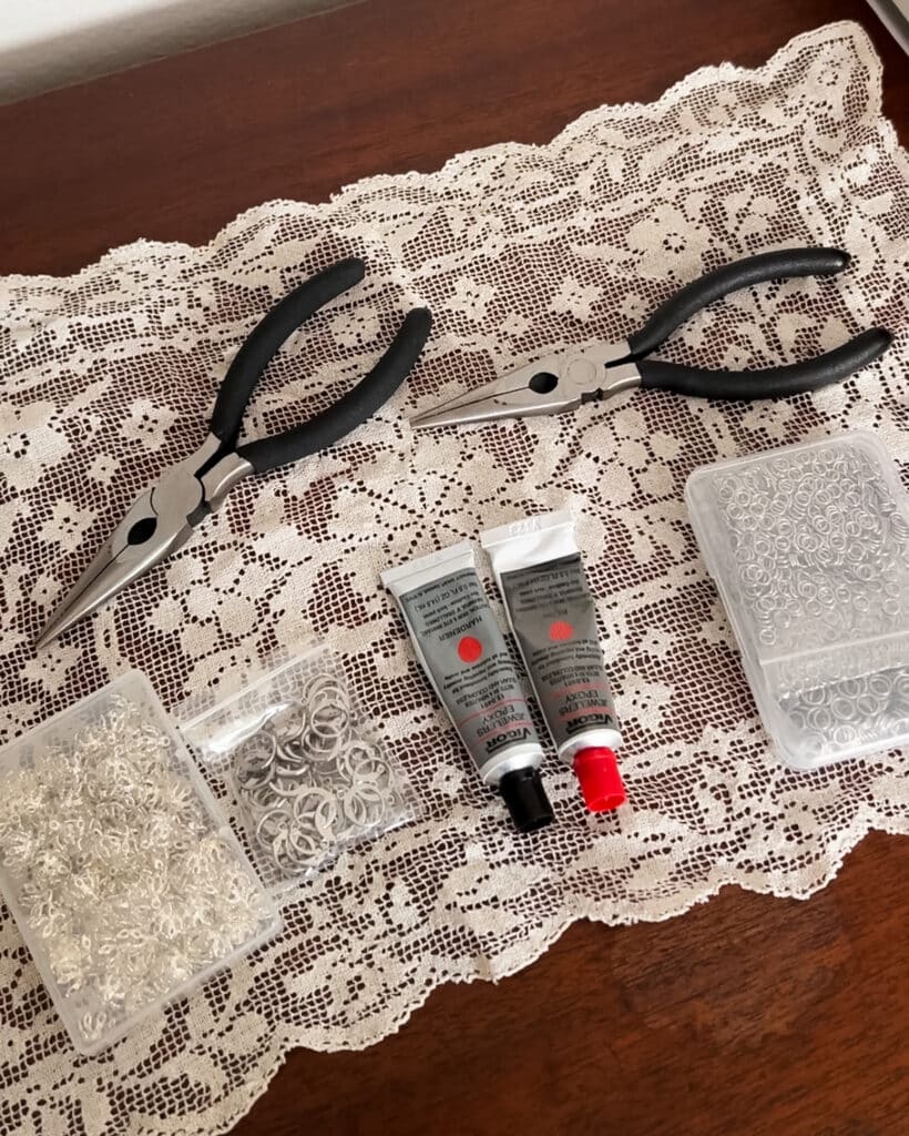 A pile of jewelry-making supplies sit on top of a white doily on a dark wood table. They include bead caps, earring findings, two tubes of epoxy, a package of jump rings, and two sets of needle nose pliers.