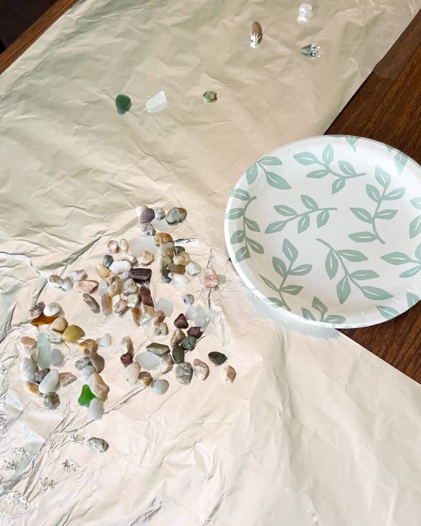 A few pieces of aluminum foil are spread on top of a table and covered with beach pebbles and sea glass. A paper plate for mixing epoxy sits nearby.