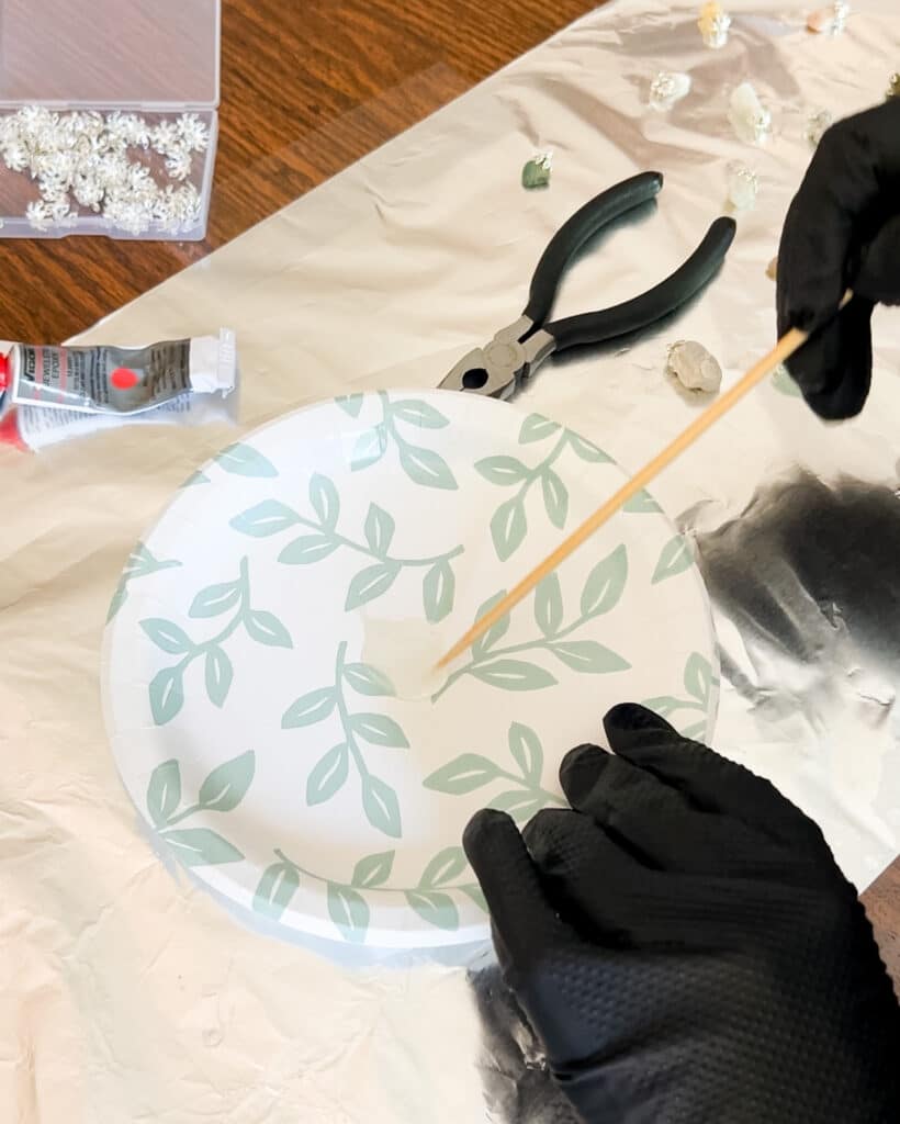 A person wearing black nitrile gloves mixes jewelers epoxy on a paper plate using a bamboo skewer.