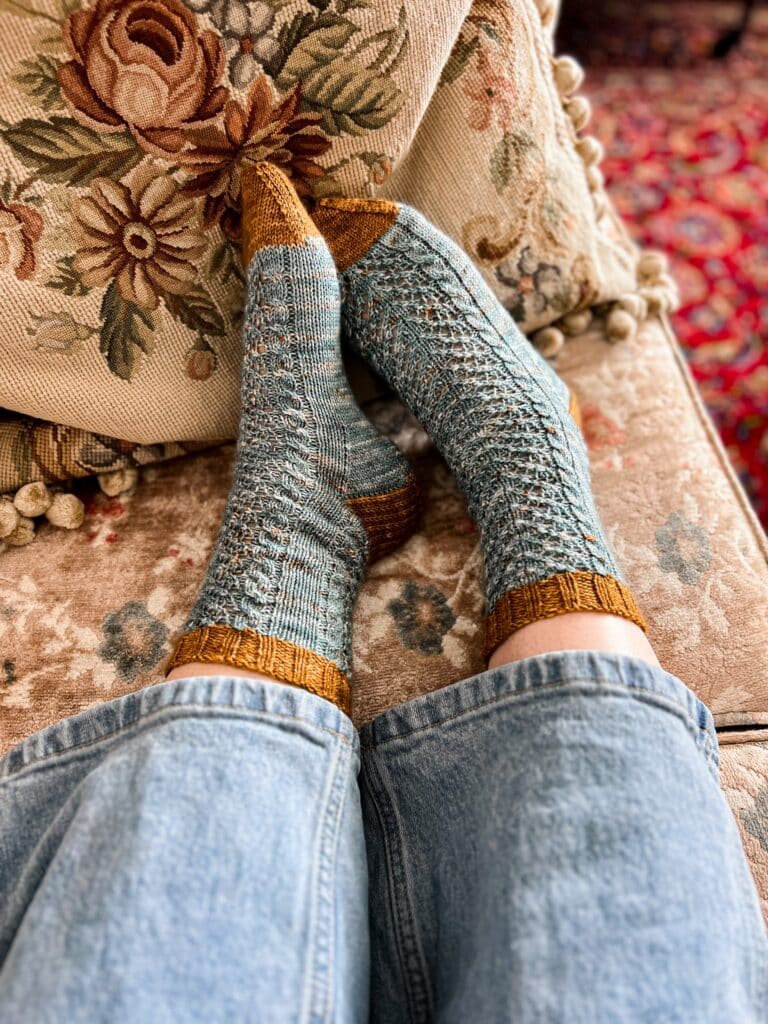A woman's feet wearing handknit socks rest against a needlepoint cushion. The socks are a speckled teal with contrasting brown cuffs, heels, and toes.