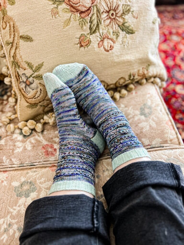 A woman's feet wearing handknit socks rest against a needlepoint cushion. The socks are a heavily variegated mix of blues and greens with contrasting aqua cuffs, heels, and toes.