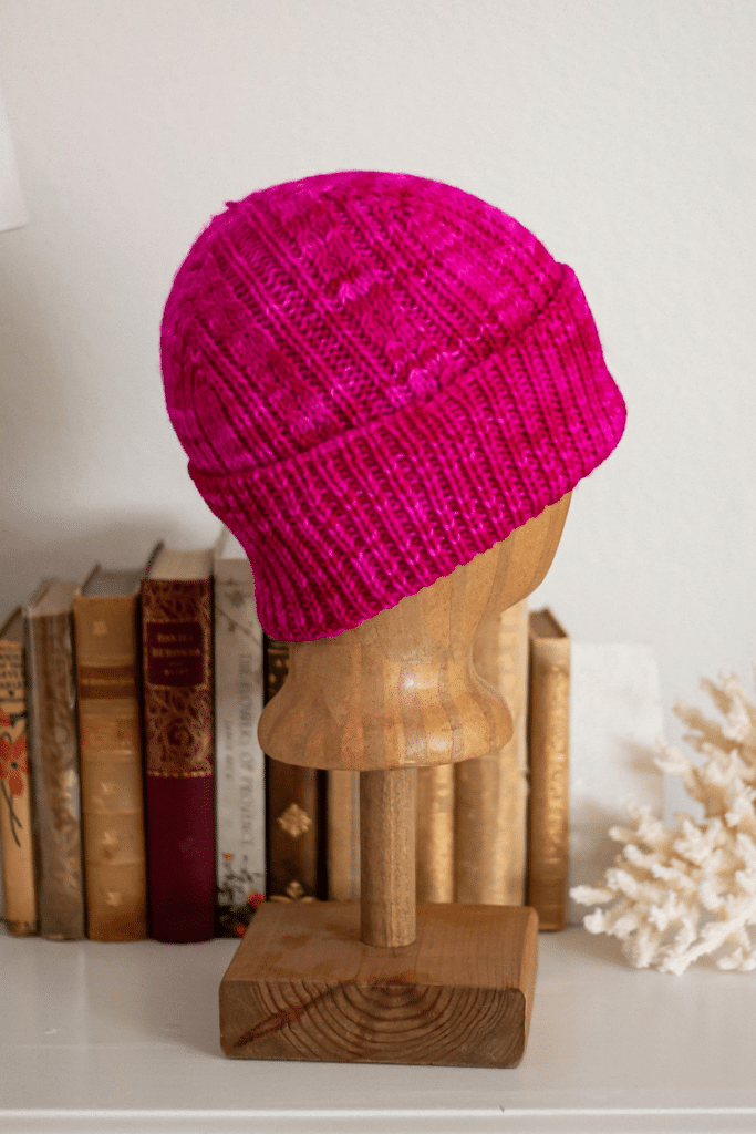 A hot pink cabled hat with a folded up brim is displayed on a wooden mannequin head in front of several antique books.