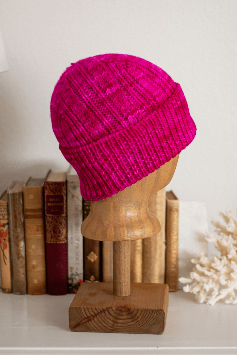 A hot pink cabled hat with a folded up brim is displayed on a wooden mannequin head in front of several antique books.