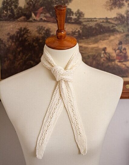 A white, lacy scarflet is tied around the neck of a white dressmaker's form. The ends of the scarflet hang down over the chest of the dressmaker's form.