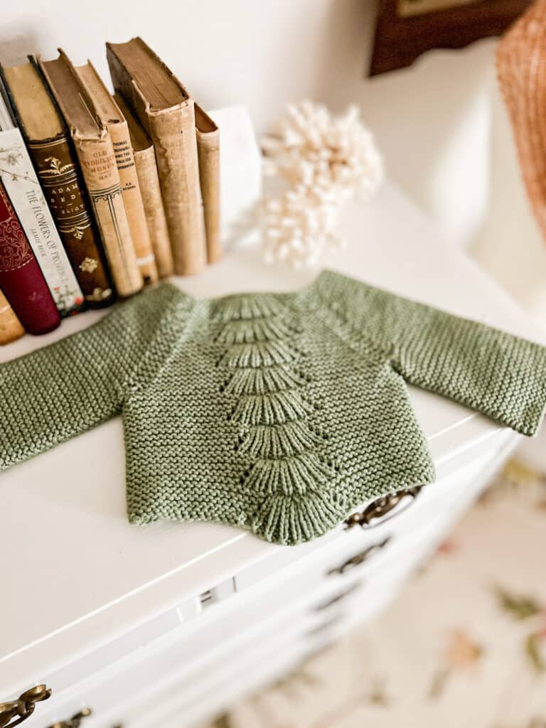 A small, green, handknit baby sweater with a garter stitch body and lacy panel down the front sits on top of a white dresser with lots of antique books in the background.