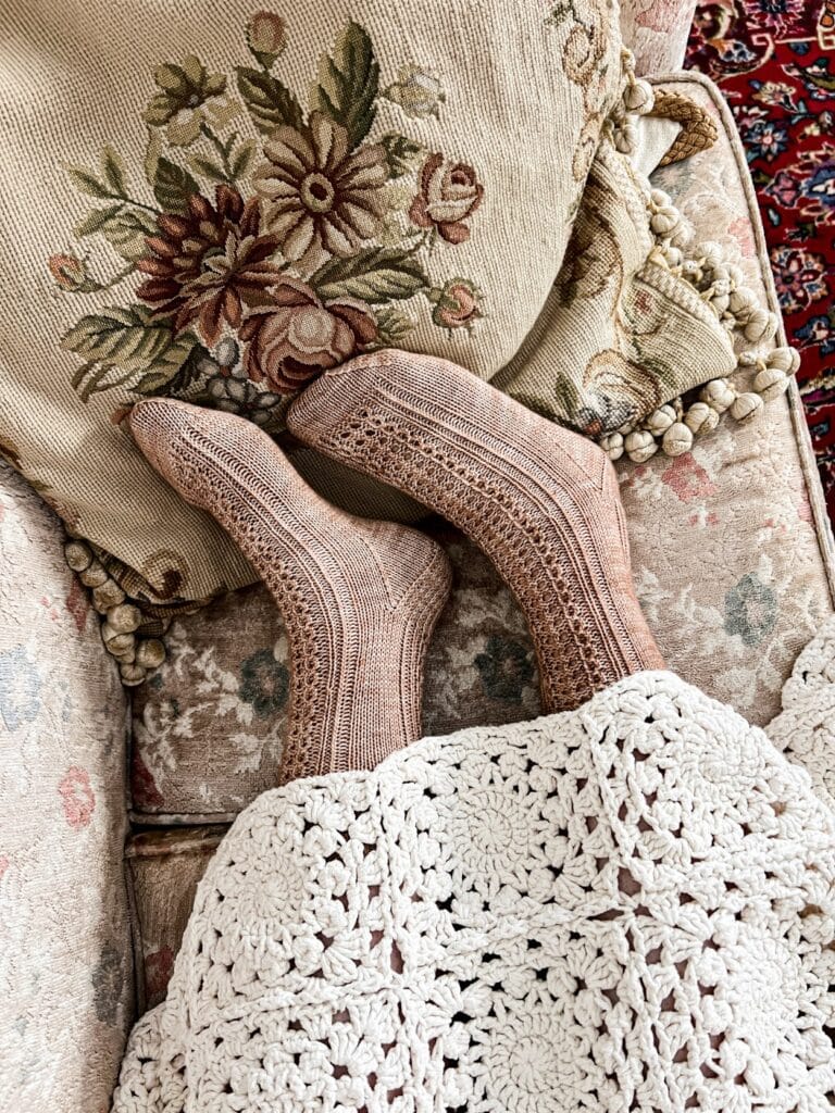 A woman's feet protrude from under a white crochet blanket with some handknit socks on them. The socks are knit in a dusty pink clay color and feature panels of lacy eyelets down the front and back.