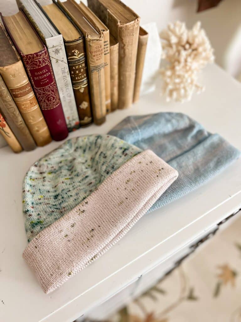 Two Mussleburgh Hats with folded brims, one in pink and green, one in a subtly variegated blue, sit on top of a white dresser with their sides overlapping. There are lots of antique books in the background.