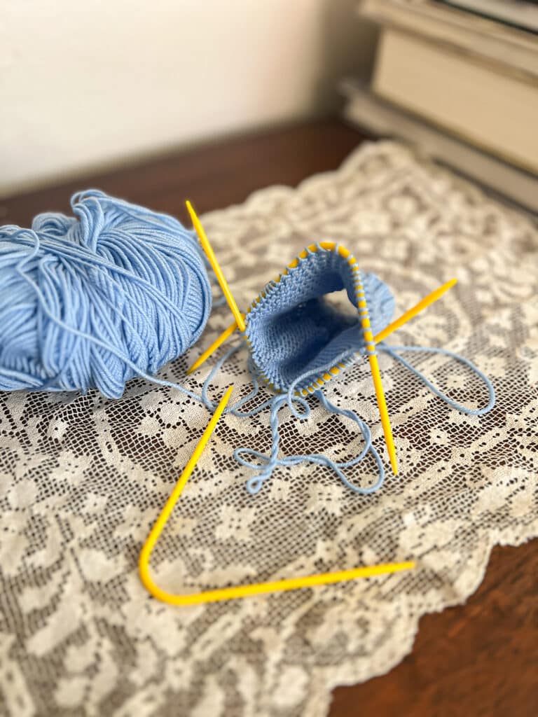 A light blue knit sock in progress on a pair of bent yellow plastic needles rests on a doily next to a third yellow bent needle.