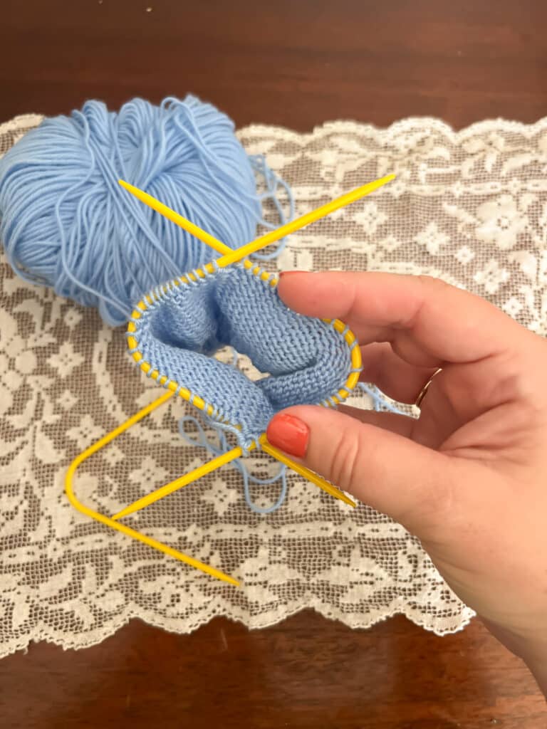 A white woman's right hand holds a light blue knit sock in progress on a pair of yellow plastic knitting needles with a rigid curve in the middle of each needle.