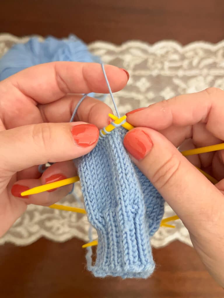 A white woman wearing coral-colored nail polish knits a light blue sock on yellow plastic knitting needles.