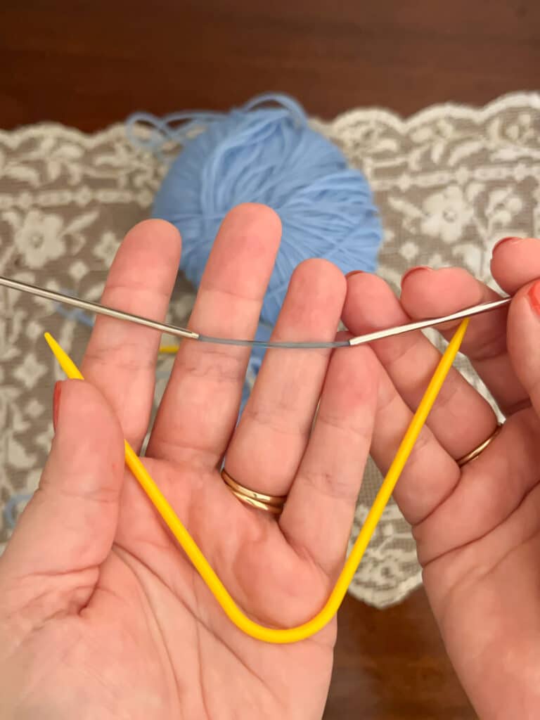 A photo showing a white woman's hands holding two types of double-pointed needles. One is yellow plastic and permanently curved at about a 45-degree angle. The other has metal tips with a very small length of blue cord connecting them. The hand holding that needle is letting the needle relax so that the needle is almost completely straight, which the rigid plastic needle cannot do.