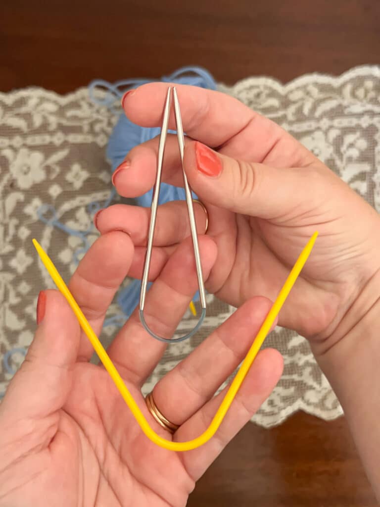 A photo showing a white woman's hands holding two types of double-pointed needles. One is yellow plastic and permanently curved at about a 45-degree angle. The other has metal tips with a very small length of blue cord connecting them. The hand holding that needle is bending it so that the needle tips are touching, which the rigid plastic needle cannot do.