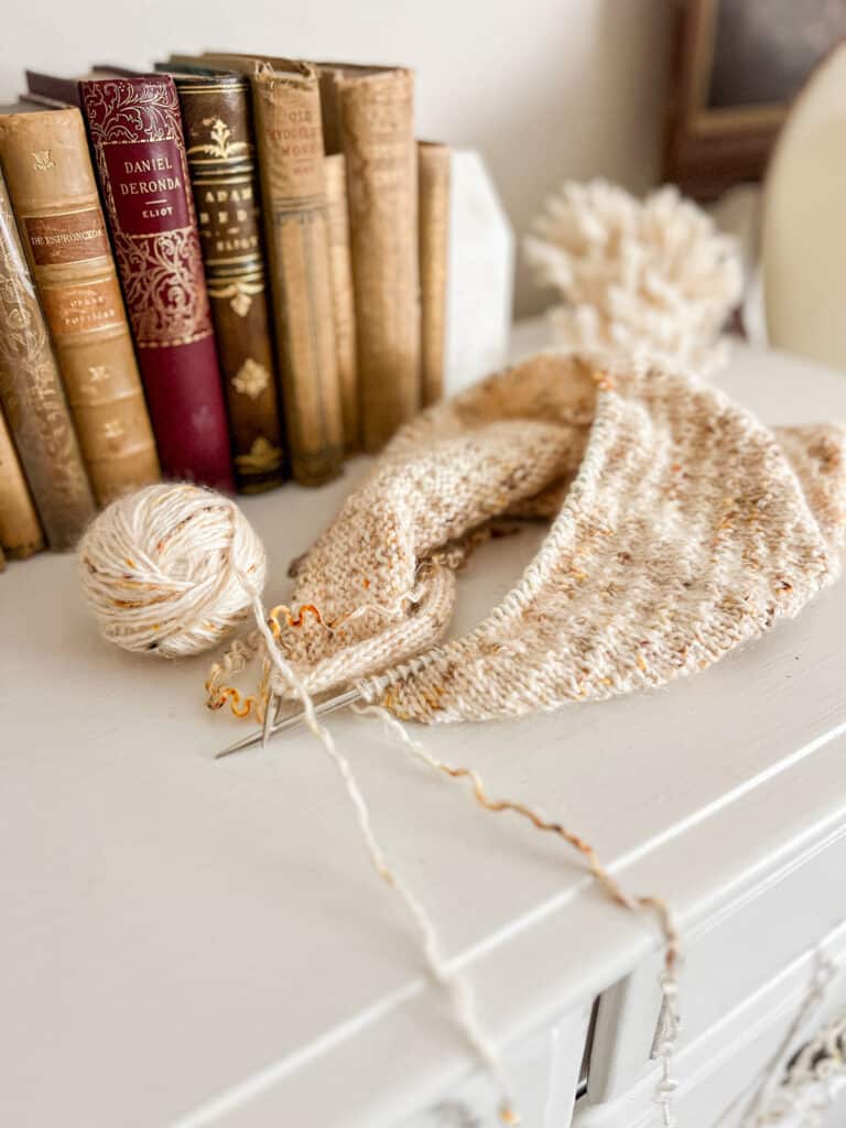 A white knit in progress with orange and brown speckles sits on top of a white dresser with lots of antique books in the background.