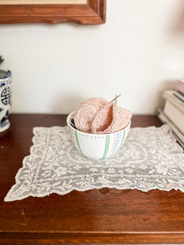 A pastel knit sock in progress sits in a ceramic bowl with painted pastel decorations on top of a vintage white doily.