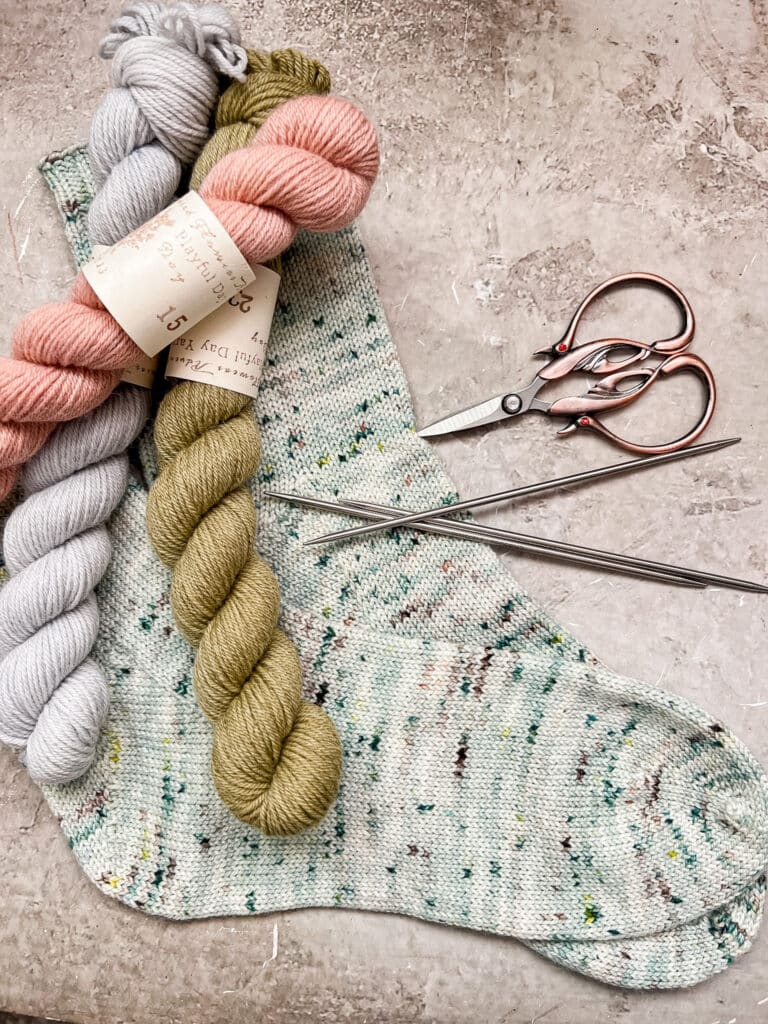 A pair of green handknit socks sits on a gray speckled surface along with three mini skeins of yarn, some double-pointed needles, and a pair of scissors.
