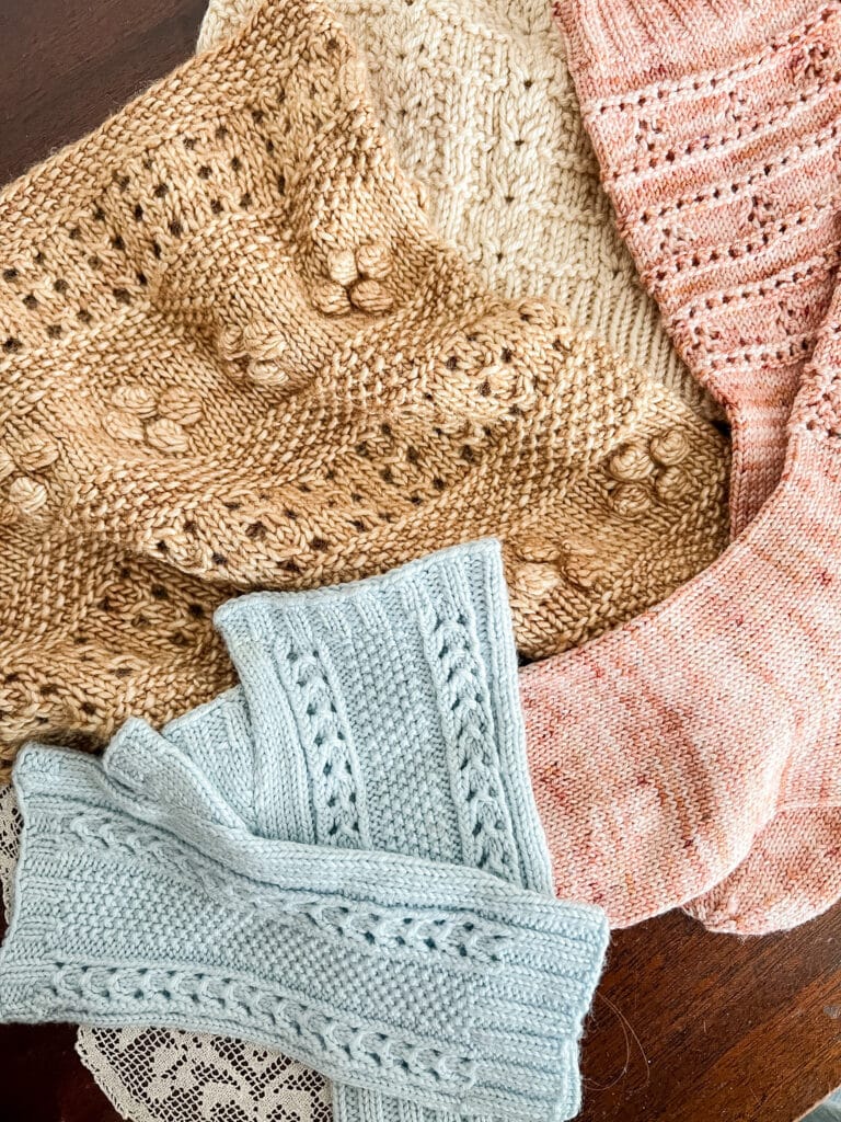 A pile of pastel knits in shades of pink, blue, and tan sit on top of a table. The knits are full of texture including eyelets, twisted stitches, and bobbles.