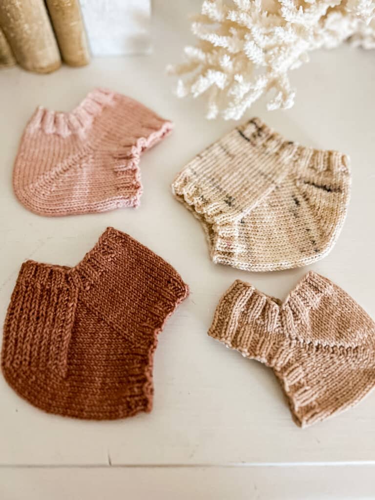Four different knit sock heels (fish lips kiss heel, Fleegle heel, German short row heel, and heel flap and gusset) are laid out flat on top of a white dresser.