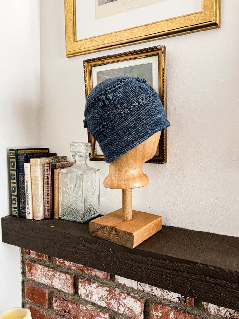 A dark blue knit hat with a folded brim and bobble flowers around the body is displayed on a brown wooden head form. The form sits on a dark brown mantel next to several antique books and in front of some antique prints in gilded frames.