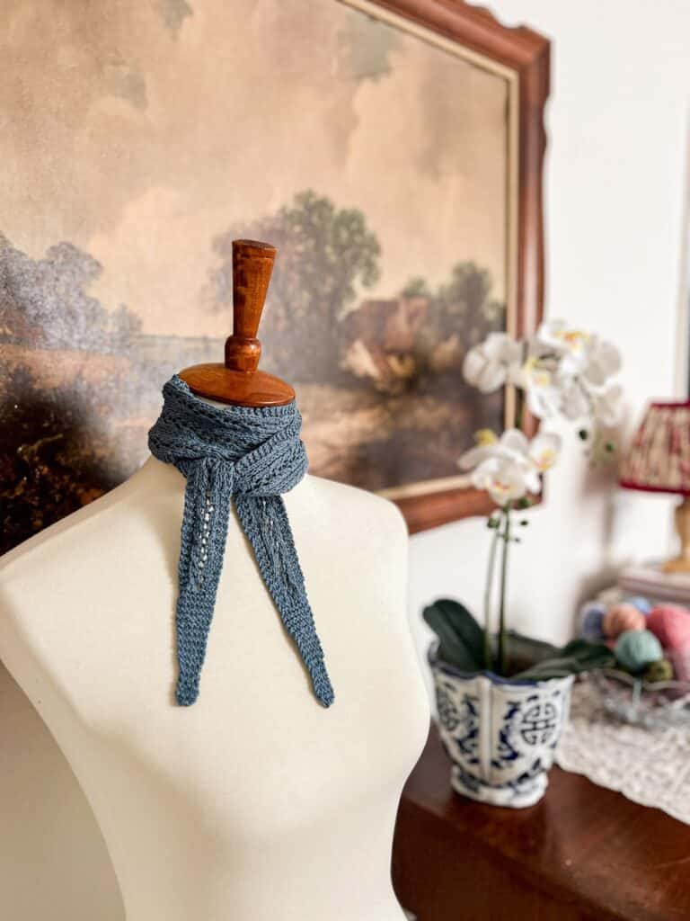 A small, blue scarf with lacy center and garter-stitch edges is tied around the neck of a white dressmaker's form. In the background are a print of a large landscape painting and a dark wood table with some orchids and a bowl of yarn balls on it.